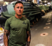 An Australian Army soldier with tattoos standing infront of military vehicles wearing a tactical green shirt with logo of Odins horns on the chest