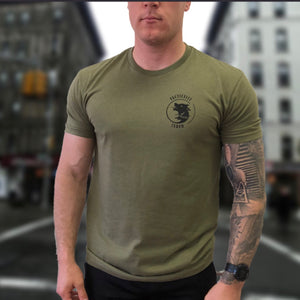 a tattooed man wears a military green undershirt and a black watch walking down the street