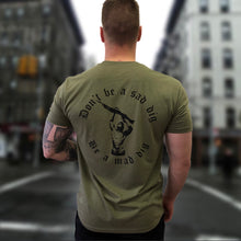 a soldier stands in the street with a green undershirt. the shirt has text and a picture of a man holding a heavy rifle with writing saying don't be a sad dig, be a mad dig